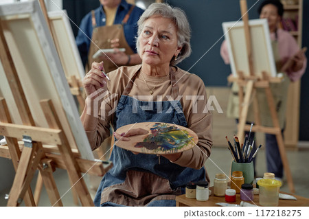 Waist up portrait of creative Caucasian senior woman painting picture on easel and wearing apron while enjoying hobby in art studio copy space Waist up portrait of creative Caucasian senior woman painting picture on easel and wearing apron while enjoying hobby in art studio copy space 117218275