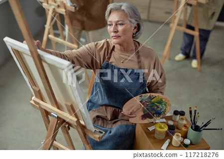 High angle portrait of creative mature woman painting picture on easel in art studio and holding palette pursuing inspiration 117218276