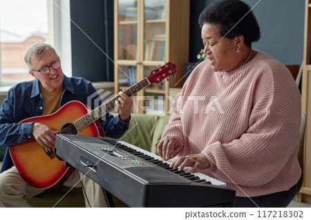 Side view portrait of Black senior woman playing piano and singing with senior man with guitar in background copy space Side view portrait of Black senior woman playing piano and singing with senior man with guitar in background copy space 117218302
