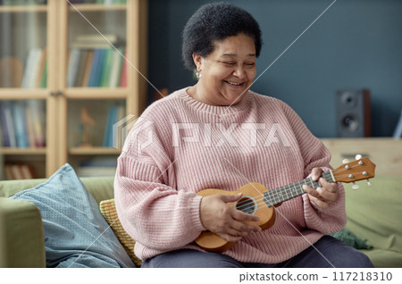 Portrait of happy senior woman playing ukulele at home and enjoying creative hobby in retirement copy space 117218310
