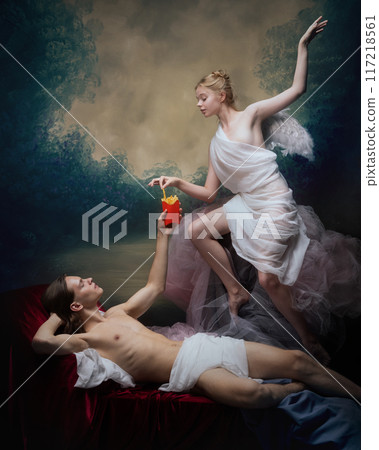 Man taking fries from graceful woman with small wings, both in classical drapery against vintage background. Blend of modern and classical elements. Earthly desires 117218561