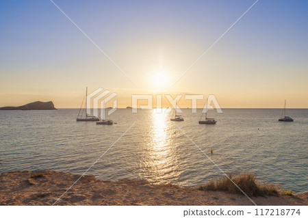 Cala Conta beach at sunset in Ibiza sailboats anchored in tranquil sea warm sun illuminating horizon Cala Conta beach at sunset in Ibiza sailboats anchored in tranquil sea warm sun illuminating horizon 117218774