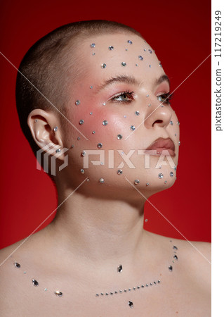 Vertical portrait of bald young woman wearing make up with rhinestones and looking away on red 117219249