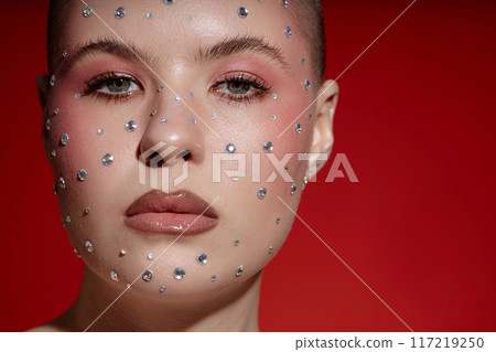 Cropped closeup portrait of confident woman wearing creative makeup with rhinestones and looking at camera over red, copy space Cropped closeup portrait of confident woman wearing creative makeup with rhinestones and looking at camera over red, copy space 117219250