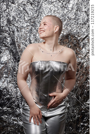Vertical fashion portrait of young woman wearing silver outfit posing with smile over metallic background 117219255