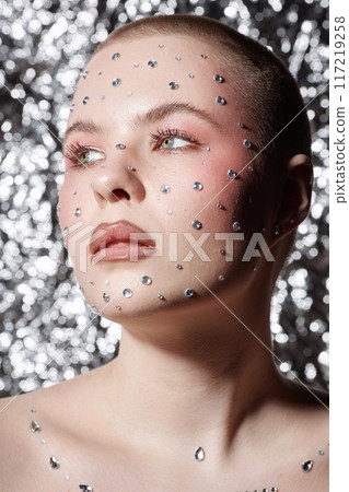 Vertical close up portrait of bald young woman wearing make up with silver gems looking away on metallic background Vertical close up portrait of bald young woman wearing make up with silver gems looking away on metallic background 117219258