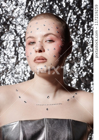 Vertical portrait of young woman wearing glittering makeup with gems looking at camera with confidence on metallic background Vertical portrait of young woman wearing glittering makeup with gems looking at camera with confidence on metallic background 117219259