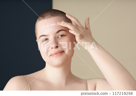 Minimal portrait of smiling young woman with no makeup applying face cream and looking at camera on two tone background Minimal portrait of smiling young woman with no makeup applying face cream and looking at camera on two tone background 117219298