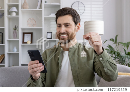 Smiling man holding new house keys while looking at phone. Depicts excitement, real estate success, new home ownership, and modern lifestyle. Concept of achievement and digital communication. 117219859