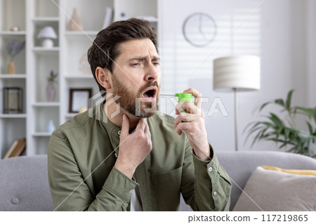 Man using throat spray for sore throat relief sitting on sofa. Demonstrates home remedies, health and wellness. Expression of discomfort and need for medication. Indoor setting, casual attire. Man using throat spray for sore throat relief sitting on sofa. Demonstrates home remedies, health and wellness. Expression of discomfort and need for medication. Indoor setting, casual attire. 117219865