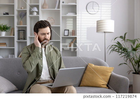 Man sitting on sofa using laptop with phone and experiencing ear pain. Conceptual image of discomfort, health issues, and technology. Man sitting on sofa using laptop with phone and experiencing ear pain. Conceptual image of discomfort, health issues, and technology. 117219874