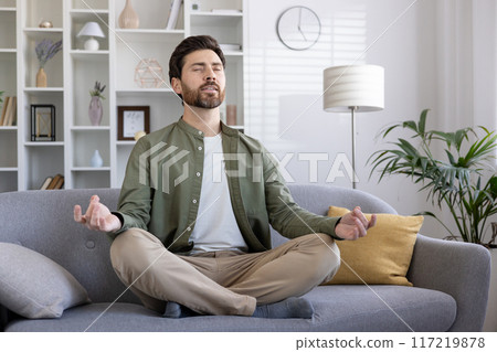 Man meditating on sofa in living room, practicing mindfulness and achieving relaxation. Person maintaining calm and inner peace at home with eyes closed in meditative pose Man meditating on sofa in living room, practicing mindfulness and achieving relaxation. Person maintaining calm and inner peace at home with eyes closed in meditative pose 117219878