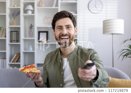 Cheerful man holding slice of pizza while watching TV at home. Smiling adult relaxing on sofa with remote control and delicious snack, enjoying leisure time 117219879