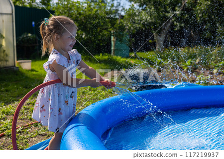 A child in a dress splashes water and laughs while filling the pool. In summer, the child fills the pool with water and splashes from the hose. Cute child enjoys summer and water. A child in a dress splashes water and laughs while filling the pool. In summer, the child fills the pool with water and splashes from the hose. Cute child enjoys summer and water. 117219937