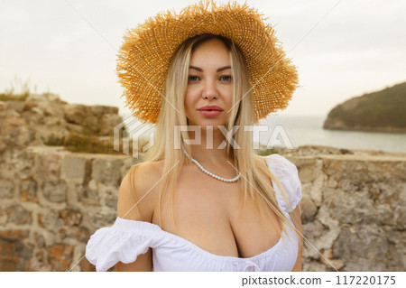 Charming fantasy woman in a hat posing for the camera. Young spectacular woman in a summer dress with bare shoulders in the background on a sunny summer beach 117220175