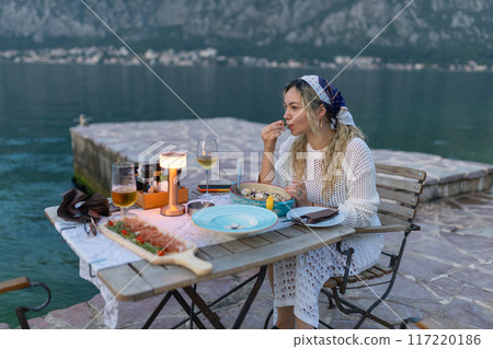A woman eats mussels, a traditional Montenegrin dish, while sitting on the terrace of a restaurant near the Bay of Kotor. Seafood and good summer vacation concept A woman eats mussels, a traditional Montenegrin dish, while sitting on the terrace of a restaurant near the Bay of Kotor. Seafood and good summer vacation concept 117220186