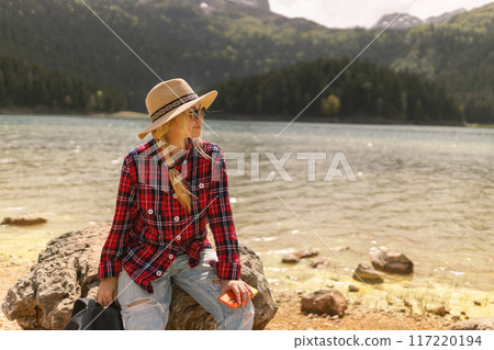 Woman traveler wearing hat and looking at amazing mountains and lake, wanderlust travel concept. black lake Montenegro. Woman traveler wearing hat and looking at amazing mountains and lake, wanderlust travel concept. black lake Montenegro. 117220194