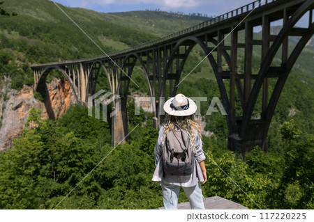 A girl in a hat stands against the backdrop of a beautiful arched bridge over the Tara River, a popular tourist attraction in Montenegro. 117220225