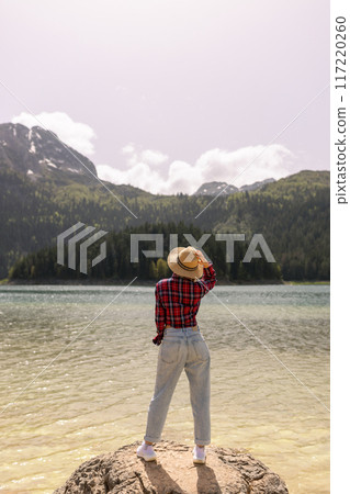Woman traveler wearing hat and looking at amazing mountains and lake, wanderlust travel concept. black lake Montenegro. Woman traveler wearing hat and looking at amazing mountains and lake, wanderlust travel concept. black lake Montenegro. 117220260