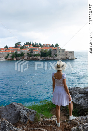 girl tourist in a hat looks from a top on the island of Sveti Stefan in Montenegro and the sea girl tourist in a hat looks from a top on the island of Sveti Stefan in Montenegro and the sea 117220277