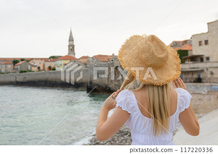 Caucasian woman wearing a straw hat stands on an observation deck looking at the old town of Budva, Montenegro. Caucasian woman wearing a straw hat stands on an observation deck looking at the old town of Budva, Montenegro. 117220336