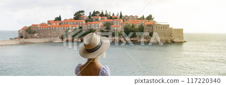 girl tourist in a hat looks from a top on the island of Sveti Stefan in Montenegro and the sea girl tourist in a hat looks from a top on the island of Sveti Stefan in Montenegro and the sea 117220340