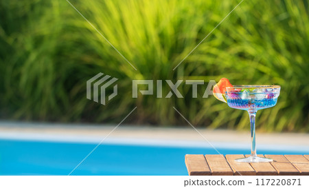 Refreshing blue cocktail with fruit garnish on a wooden table, pool and lush green background Refreshing blue cocktail with fruit garnish on a wooden table, pool and lush green background 117220871