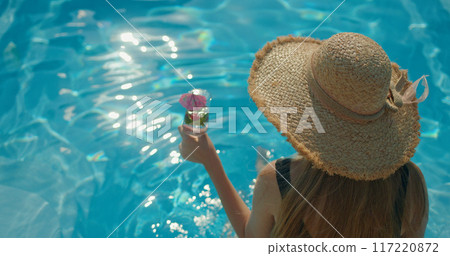 Young woman in a hat holding a cool drink, sitting on the edge of a pool on a hot summer day 117220872
