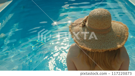 Young woman in a hat sitting by the pool, admiring the sun reflections on the water Young woman in a hat sitting by the pool, admiring the sun reflections on the water 117220873