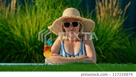 Charming middle-aged woman in a hat with a cocktail, peering out of a pool Charming middle-aged woman in a hat with a cocktail, peering out of a pool 117220874