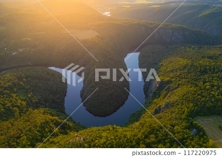 The Vltava River gracefully winds through lush greenery in Czechia during the summer, reflecting the vibrant colors of the landscape as the sun sets. 117220975