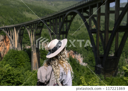 A girl in a hat stands against the backdrop of a beautiful arched bridge over the Tara River, a popular tourist attraction in Montenegro. A girl in a hat stands against the backdrop of a beautiful arched bridge over the Tara River, a popular tourist attraction in Montenegro. 117221869