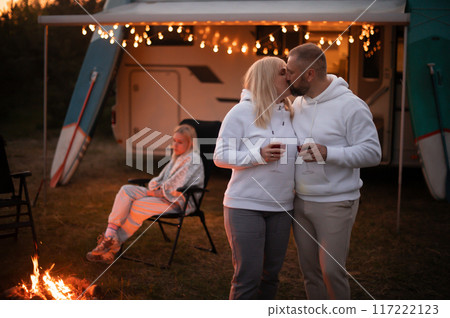 A married couple with glasses of wine stands against the background of a motorhome and rests together by the campfire. Evening family vacation 117222123