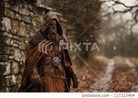 A formidable Celtic warrior clad in a hooded cloak battles the chill of winter, standing guard by a weathered stone wall as snowflakes dance around him. 117223464