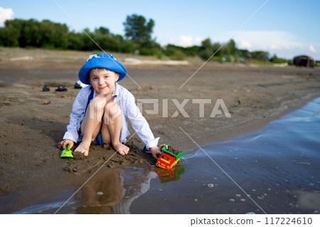 A joyful child is happily playing on the sandy beach with colorful toy trucks under the sun 117224610