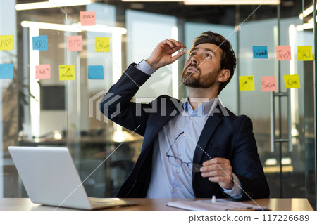 Businessman sitting at desk using eye drops in modern office. Laptop and notes on glass walls suggest busy work environment. Concept of eye strain, corporate stress relief, and workspace wellness. 117226689