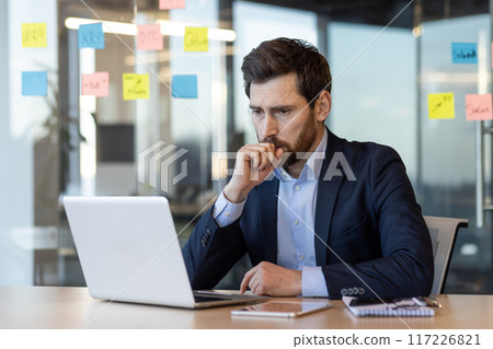 Businessman in suit working on laptop in modern office. Man appears thoughtful, reflecting, or making decisions. Background includes colorful sticky notes on glass wall, indicative of planning Businessman in suit working on laptop in modern office. Man appears thoughtful, reflecting, or making decisions. Background includes colorful sticky notes on glass wall, indicative of planning 117226821