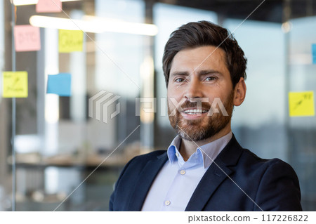 Confident businessman in professional attire smiling in office setting with sticky notes on glass wall background. Concept of leadership, success, entrepreneurship. Confident businessman in professional attire smiling in office setting with sticky notes on glass wall background. Concept of leadership, success, entrepreneurship. 117226822