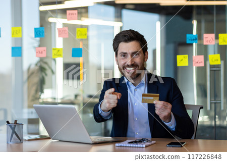 Smiling businessman sitting at office desk holding credit card. Business professional pointing towards camera, surrounded by laptop, sticky notes, and modern interior. Represents finance, success Smiling businessman sitting at office desk holding credit card. Business professional pointing towards camera, surrounded by laptop, sticky notes, and modern interior. Represents finance, success 117226848