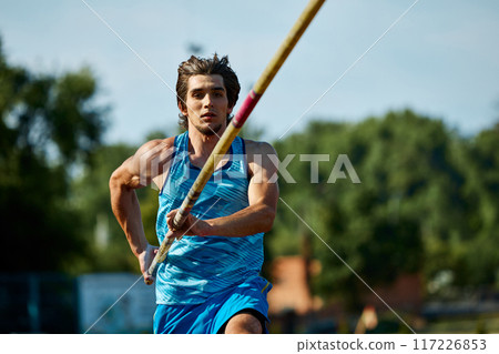 Pole vaulting athlete training on outdoor stadium track, aiming for new heights, focused muscular man running with pole, showing skills 117226853