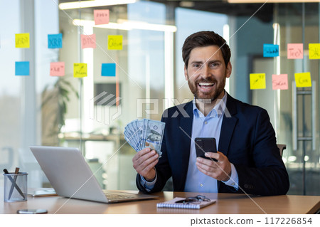 Smiling businessman holding cash and phone in modern office. Happy professional showing success with money and technology. Concept of financial achievement, technology, and workplace. 117226854