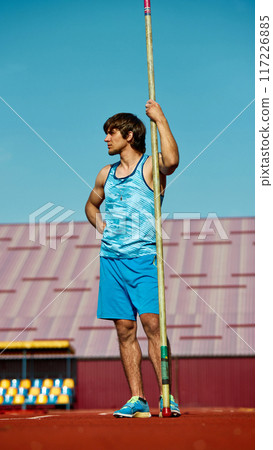 Pole vaulter in blue tank top, young athletic man standing at outdoor with pole, getting ready for intense training session under blue sky 117226885