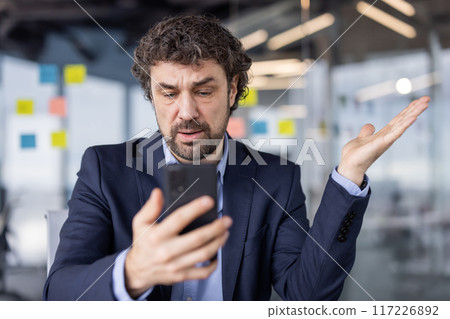 Confused businessman in suit looking at cell phone in modern office setting. Man appears perplexed or frustrated while holding smartphone, likely dealing with unexpected news or information. Confused businessman in suit looking at cell phone in modern office setting. Man appears perplexed or frustrated while holding smartphone, likely dealing with unexpected news or information. 117226892