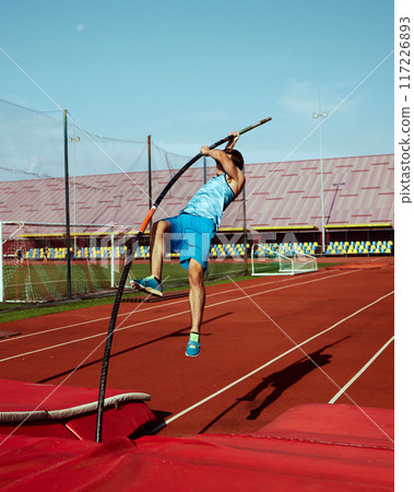 High jump practice in action, with focused and strong pole vaulter, competitive man in blue uniform training at outdoor stadium 117226893