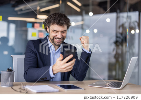 Happy businessman in suit celebrating success on smartphone in modern office. Professional expressing excitement and joy at desk with laptop, notepad, and glasses. Concept of business success Happy businessman in suit celebrating success on smartphone in modern office. Professional expressing excitement and joy at desk with laptop, notepad, and glasses. Concept of business success 117226909