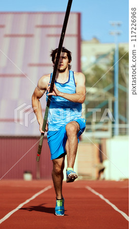 Pole jump athlete, young focused man in blue uniform on stadium track, running with pole, getting ready for high jump 117226968