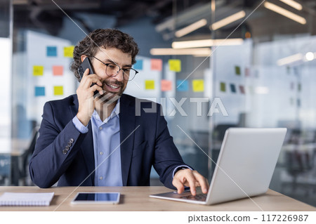 Businessman multitasking with laptop and phone in modern office. Professional in suit engaged in conversation and working on computer. Office environment with sticky notes in background. Businessman multitasking with laptop and phone in modern office. Professional in suit engaged in conversation and working on computer. Office environment with sticky notes in background. 117226987