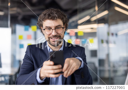 Confident businessman wearing glasses and suit using phone in modern office. Happy professional engaging with smartphone, working in contemporary business environment 117227001