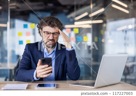 Stressed businessman holding phone with frustrated expression in modern office. Business professional dealing with work challenges. Laptop and notepad on desk, colorful notes on glass wall. 117227003