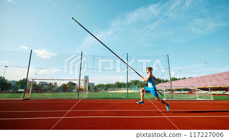 High jump training in progress, low angle view on competitive young man, pole jump athlete in motion, running with pole at outdoor track. 117227006